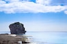 A rocky coastline with a prominent rock formation surrounded by clear blue waters and a partly cloudy sky.