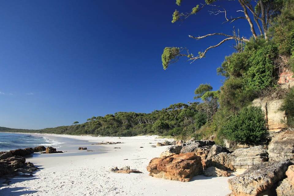 A beach with white sand, rocky outcrops, and dense green foliage.