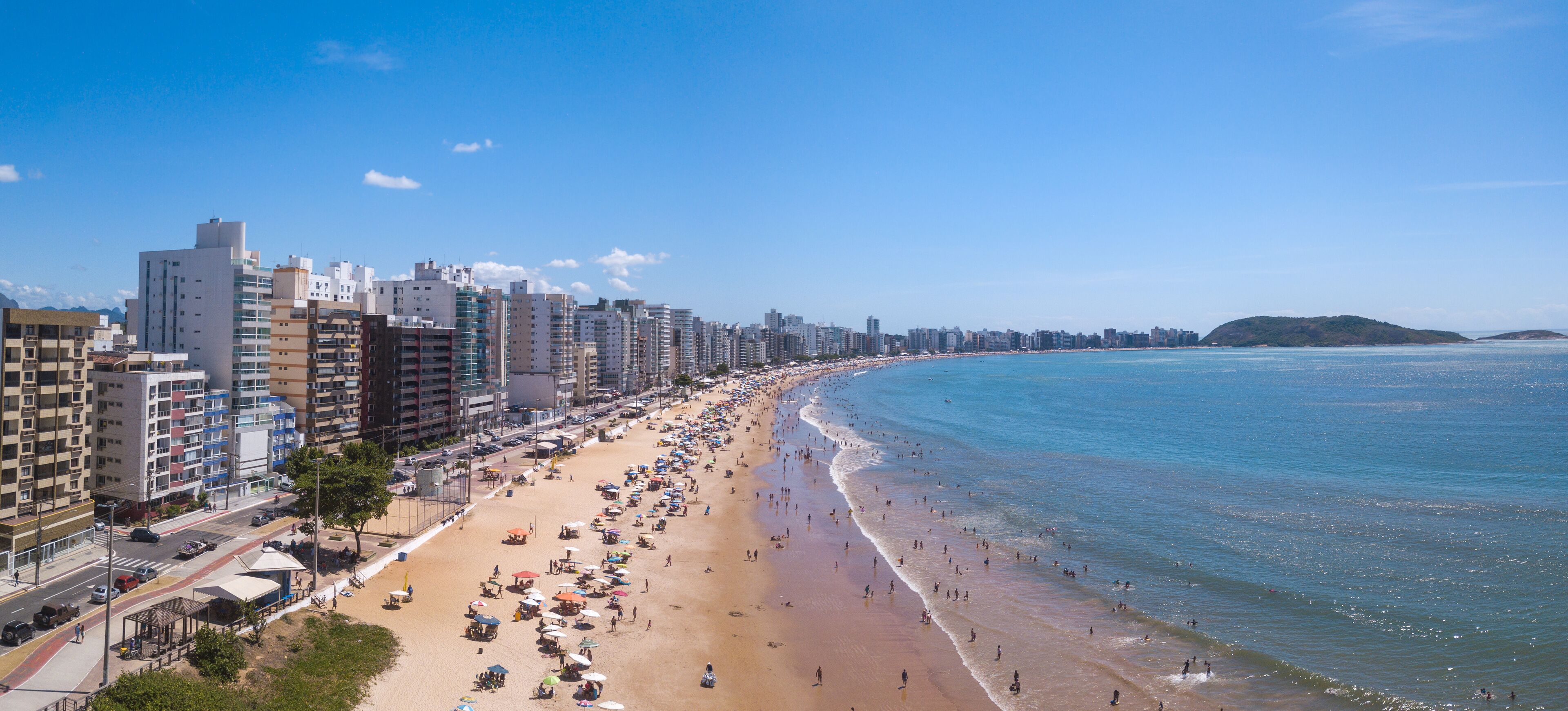 Beautiful drone view of "Praia do Morro" beach in Guarapari full of colorful umbrellas in the sand, bathers in the transparent green sea, buildings on the waterfront street and summer blue sky.