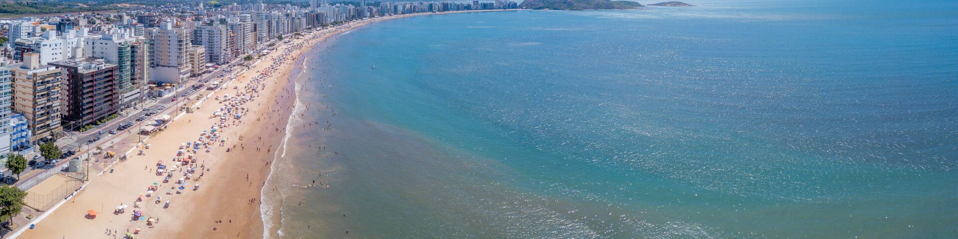 Beautiful drone view of "Praia do Morro" beach in Guarapari full of colorful umbrellas in the sand, bathers in the transparent green sea, buildings on the waterfront street and summer blue sky.