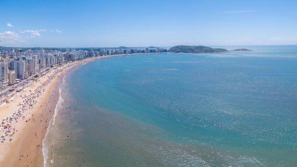 Beautiful drone view of "Praia do Morro" beach in Guarapari full of colorful umbrellas in the sand, bathers in the transparent green sea, buildings on the waterfront street and summer blue sky.
