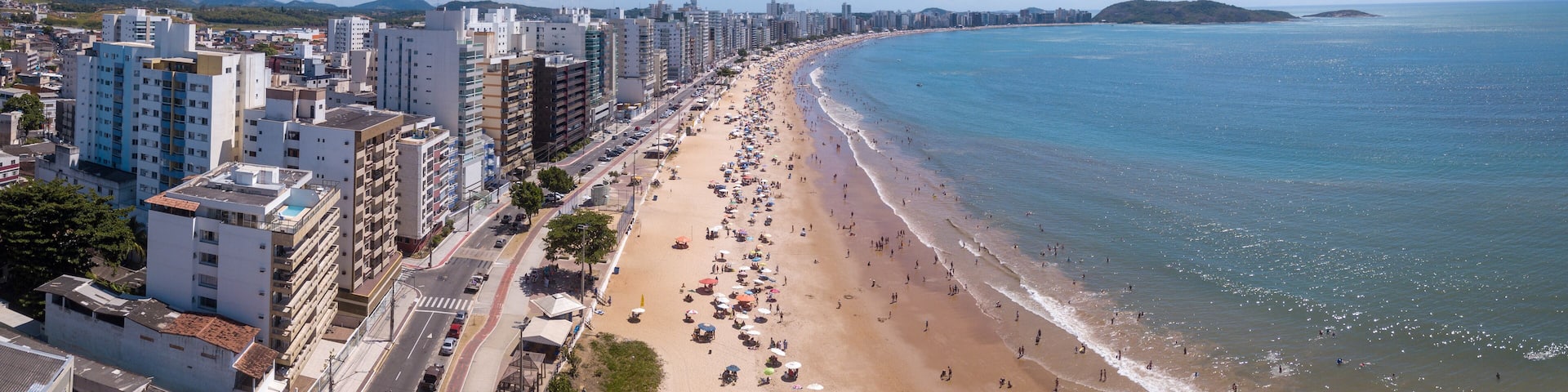 Beautiful drone view of "Praia do Morro" beach in Guarapari full of colorful umbrellas in the sand, bathers in the transparent green sea, buildings on the waterfront street and summer blue sky.