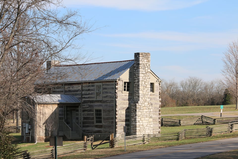 A rustic stone building with a metal roof, surrounded by a wooden fence and trees.