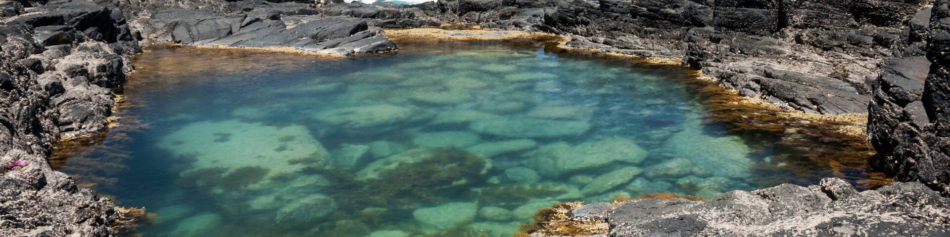 A rocky coastline with a clear pool of water, a cliff, and waves crashing against the rocks.