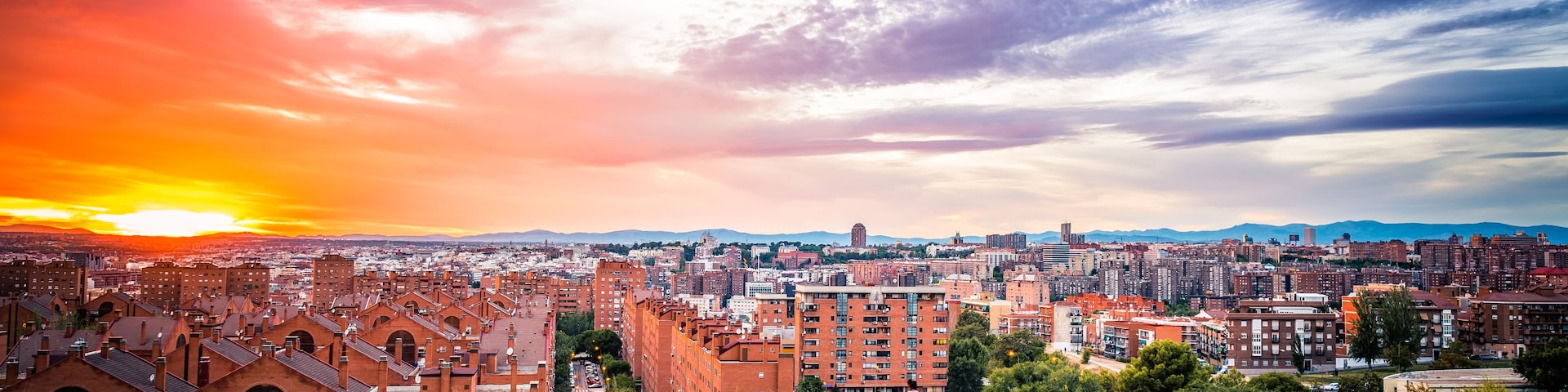 Un paisaje urbano al atardecer con un edificio de ladrillo rojo destacado en primer plano y una zona urbana densa en el fondo.