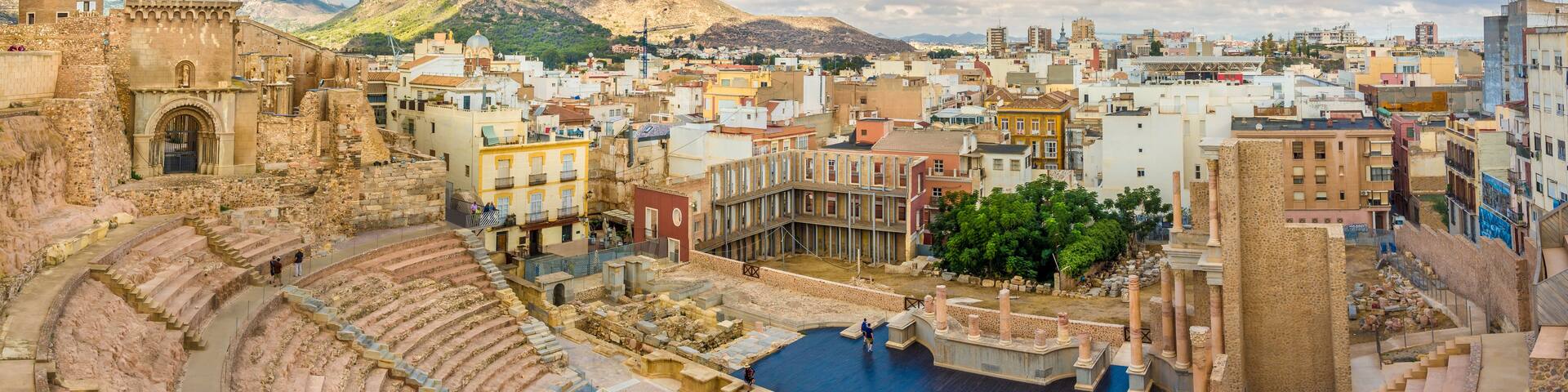 An ancient amphitheater with a pool, surrounded by historic buildings and mountains.