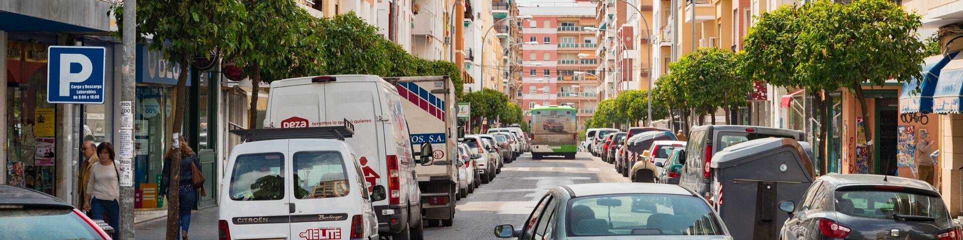 A busy urban street lined with multi-story buildings, parked cars, and a bus.