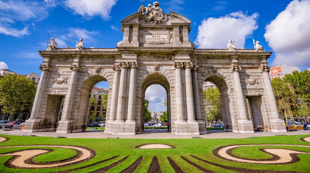 A grand archway with statues and inscriptions, surrounded by well-manicured gardens.