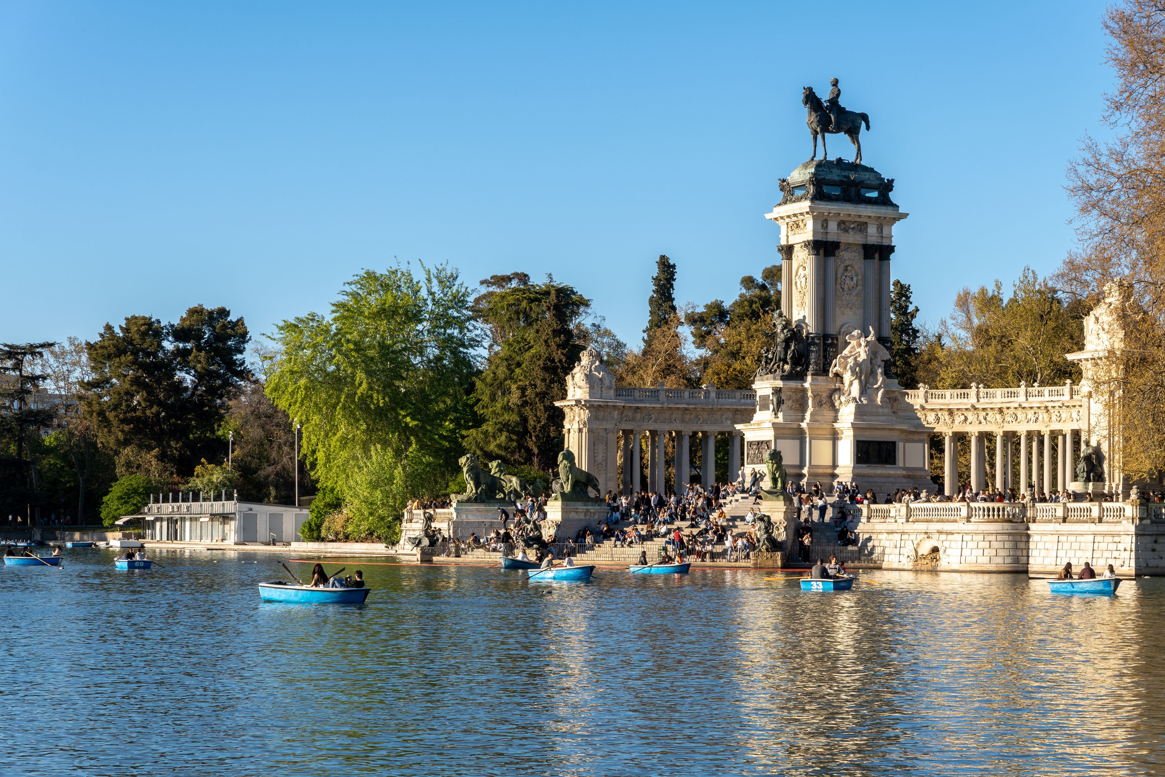 A historic fountain with statues, surrounded by trees and boats on the water.
