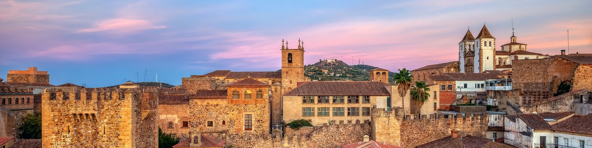 Una plaza histórica con un imponente castillo de piedra, una iglesia con campanario y una mezcla de edificios tradicionales y modernos.