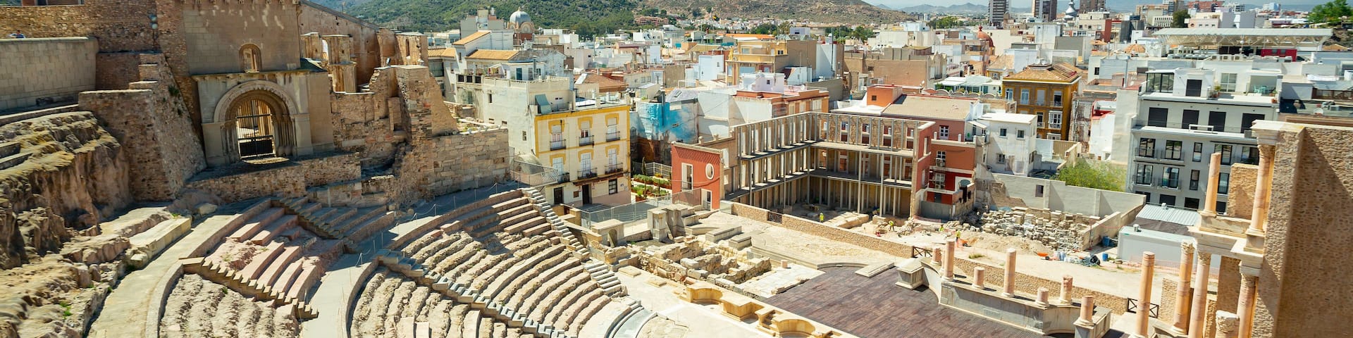 An ancient amphitheater with stone steps and a central stage, surrounded by modern buildings and a mountainous backdrop.
