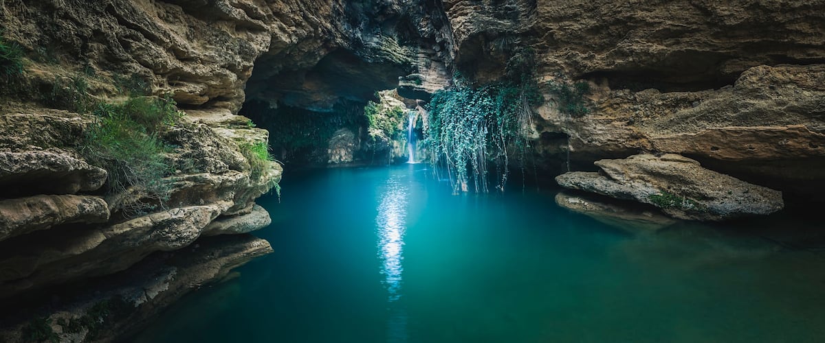 A narrow canyon with a waterfall and clear blue water.