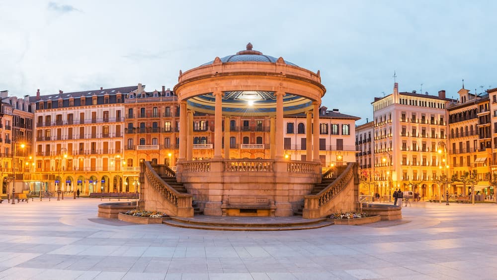 Une place historique dotée d’une fontaine centrale, entourée d’immeubles richement décorés, avec des balcons et des arcades.