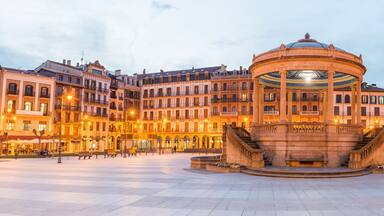 Una plaza histórica con una fuente central, rodeada de edificios ornamentados con balcones y arcos.