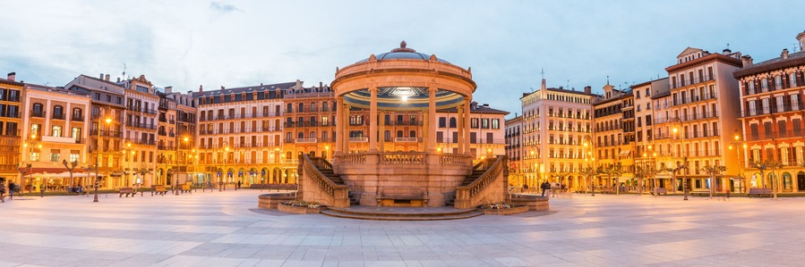 A historic plaza with a central fountain, surrounded by ornate buildings with balconies and arches.