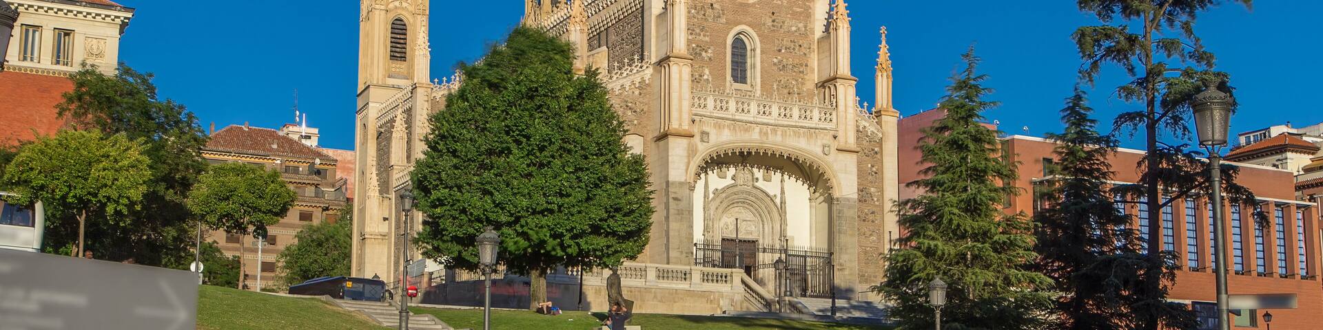 A historic cathedral with intricate architecture, a large tree in the foreground, and people relaxing on the grass.