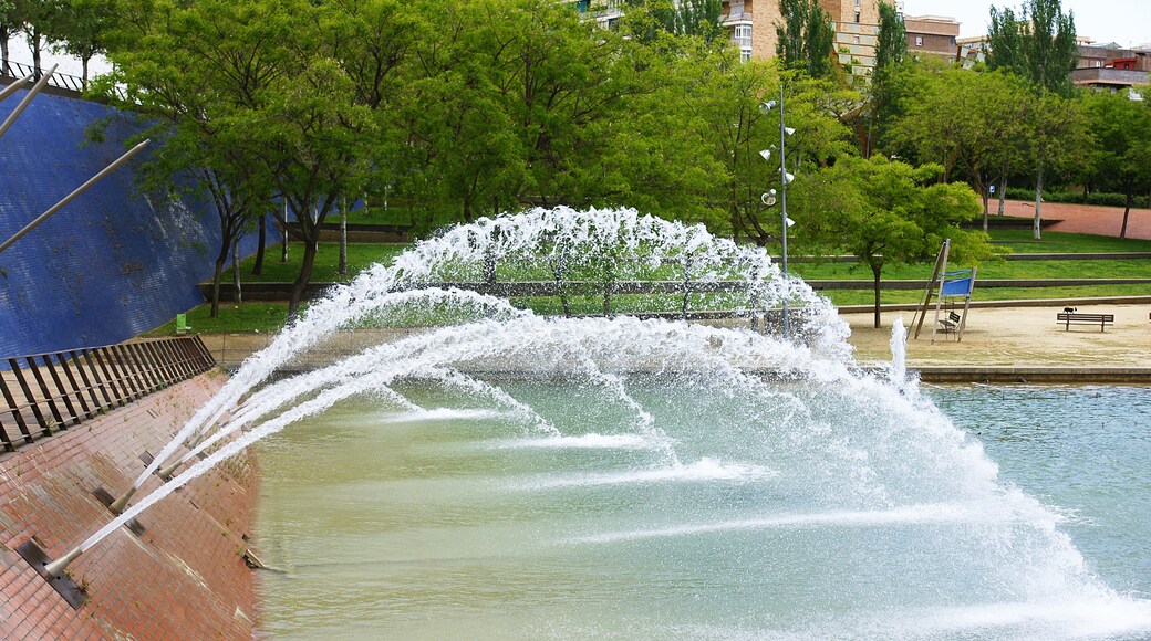 Une fontaine avec des jets d’eau, entourée d’arbres et d’un mur de briques.
