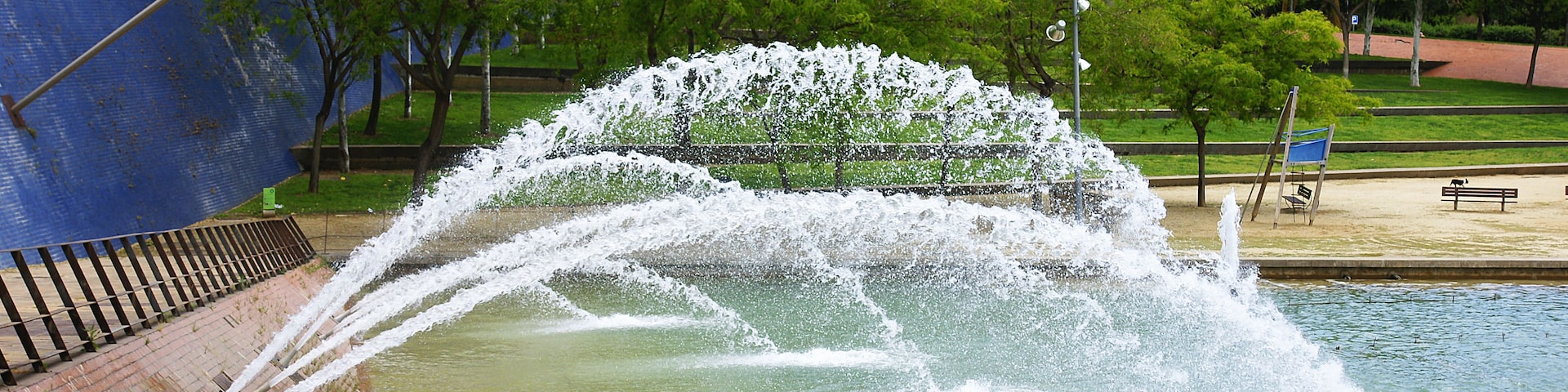 Une fontaine avec des jets d’eau, entourée d’arbres et d’un mur de briques.