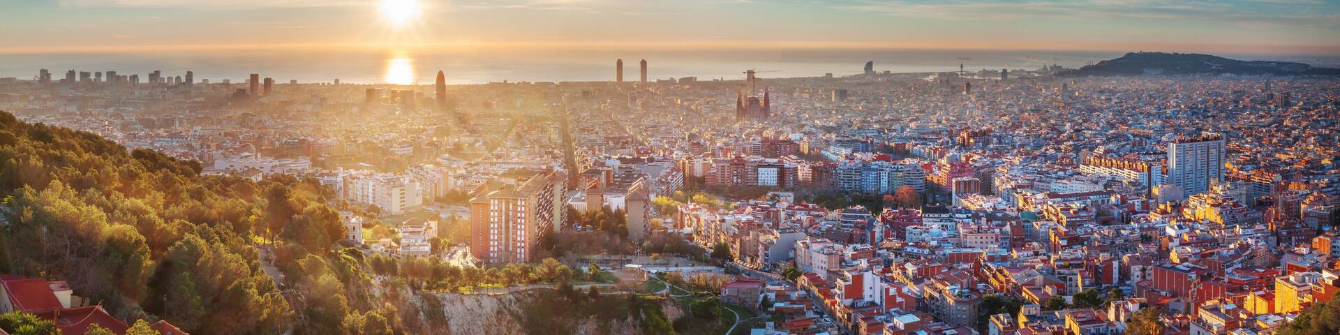 A cityscape with a dense urban area, a prominent hill in the foreground, and a clear sky with the sun setting or rising.