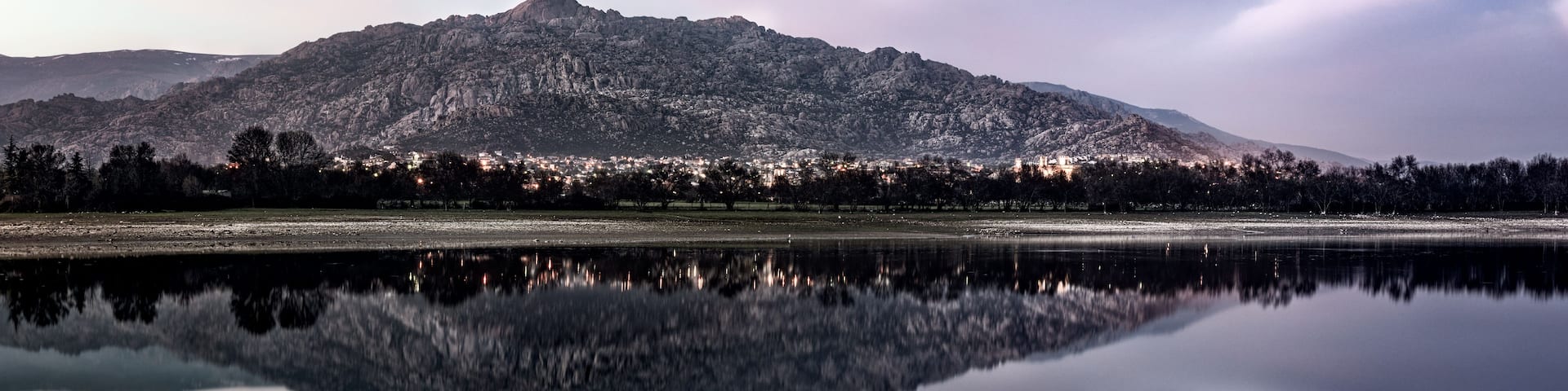 A mountain range with a lake reflecting its image, a town in the valley below, and a clear sky.