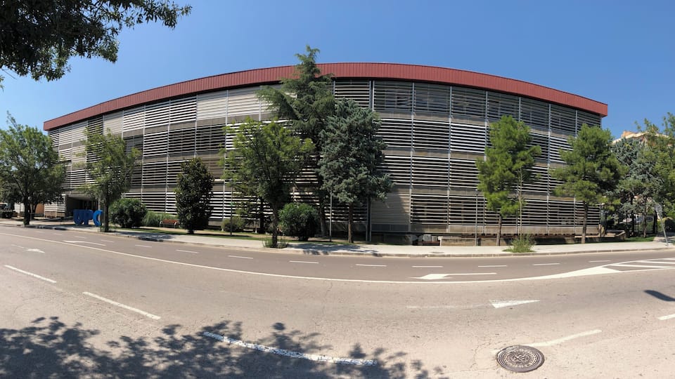 A modern building with a curved roof and horizontal slats, surrounded by trees and a clear sky.