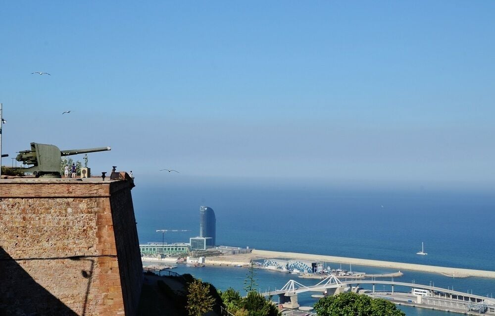 Paisatge urbà costaner amb una peça d'artilleria destacada sobre un penya-segat, un pont modern i una vista clara del mar.