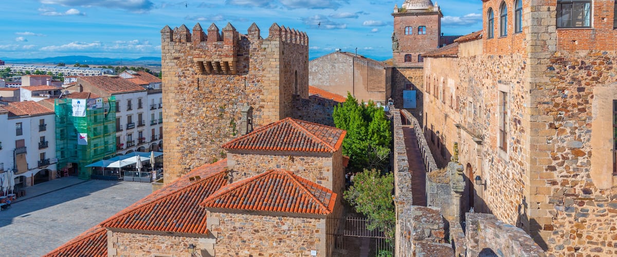 Una fortaleza de piedra histórica con tejado de tejas rojas, rodeada por una plaza adoquinada y edificios modernos.