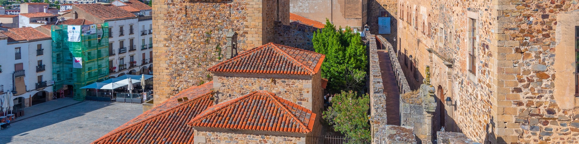 Una fortaleza de piedra histórica con tejado de tejas rojas, rodeada por una plaza adoquinada y edificios modernos.