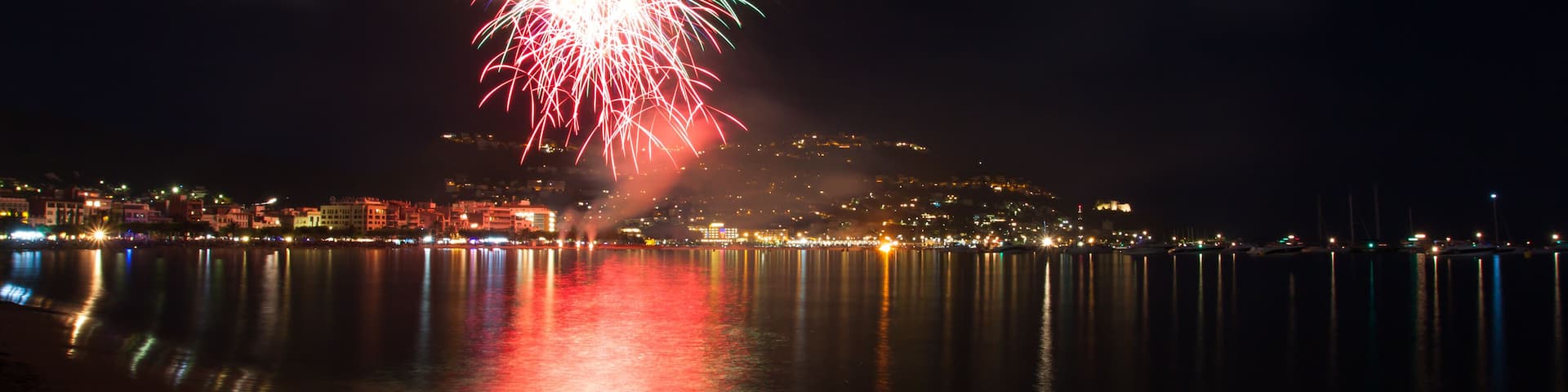 A nighttime coastal scene with a fireworks display over the water, reflecting city lights and buildings in the distance.