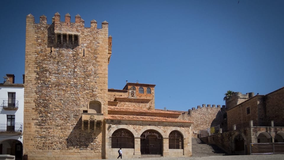 A historic stone building with a tower, arched windows, and a staircase leading to a gate.