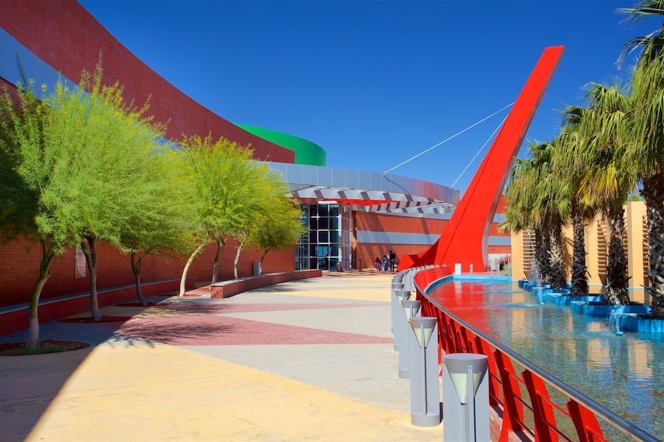 A modern building with a red and green facade, a curved glass entrance, and a water feature with palm trees.