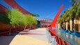 Un edificio moderno con fachada roja y verde, entrada de cristal curvo y una fuente con palmeras.
