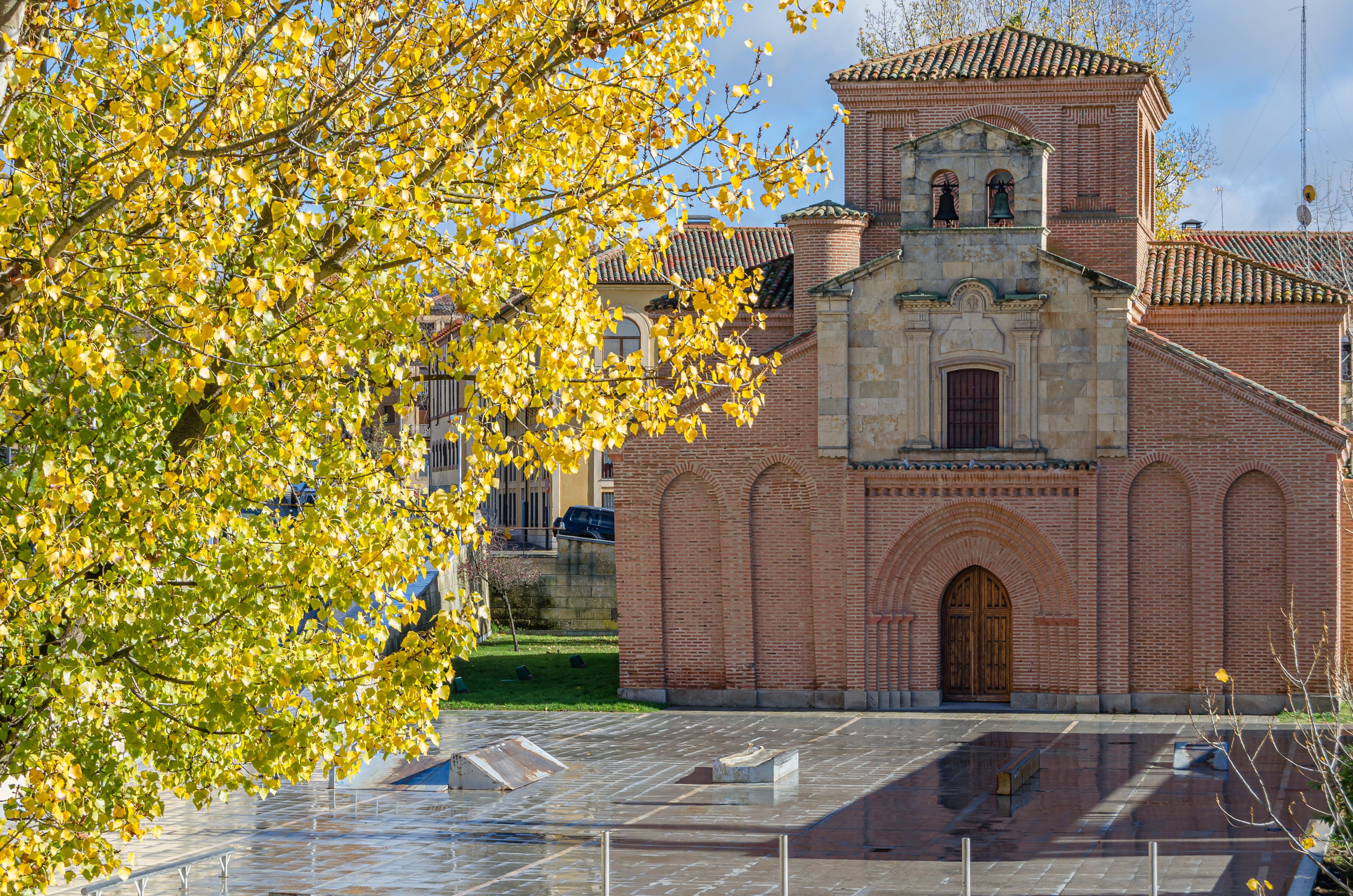 A historic church with a bell tower, surrounded by a plaza with a fountain and a tree with yellow leaves.