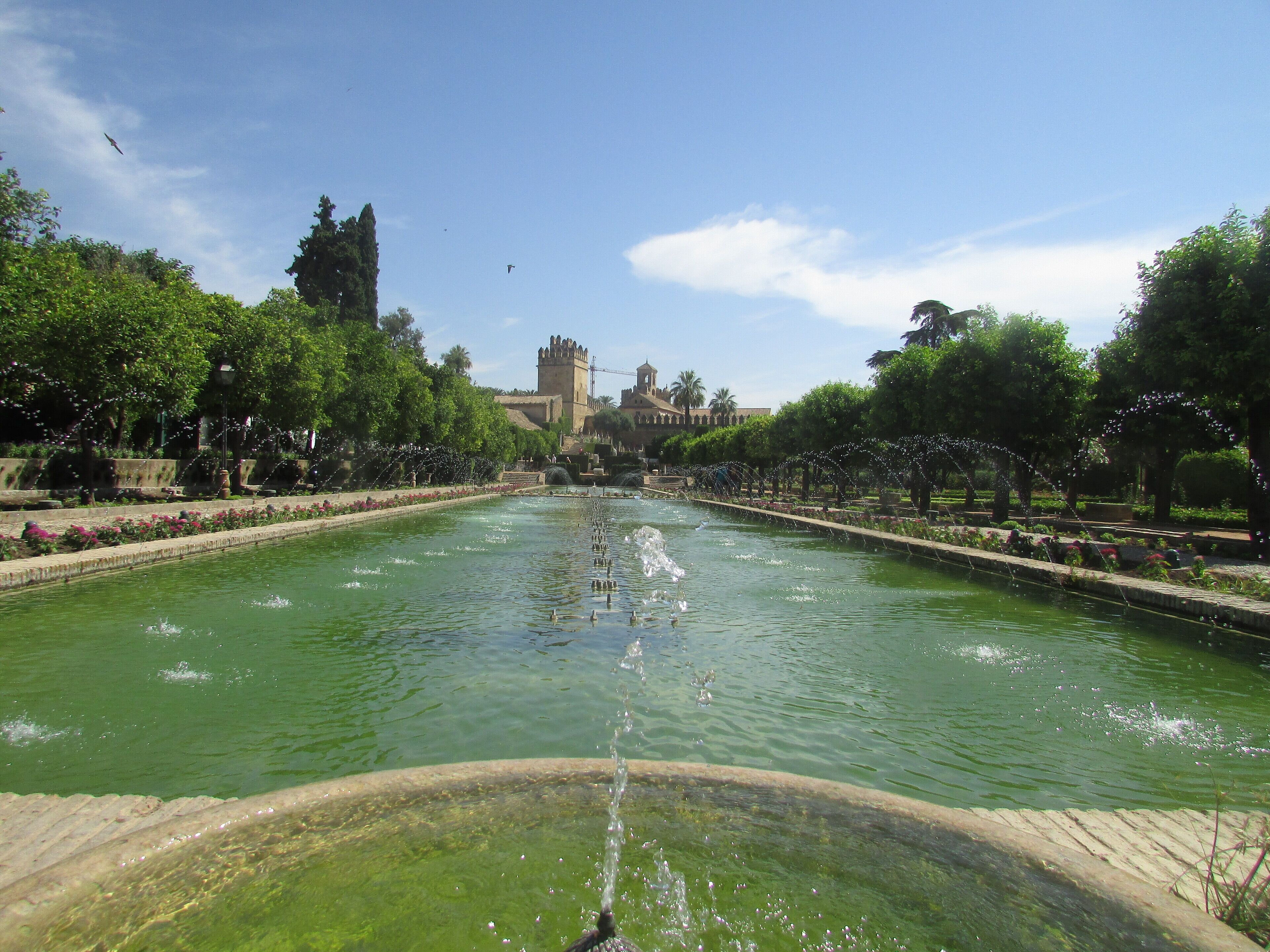 A large fountain with water jets in the center of a park, surrounded by trees and a distant castle-like building.