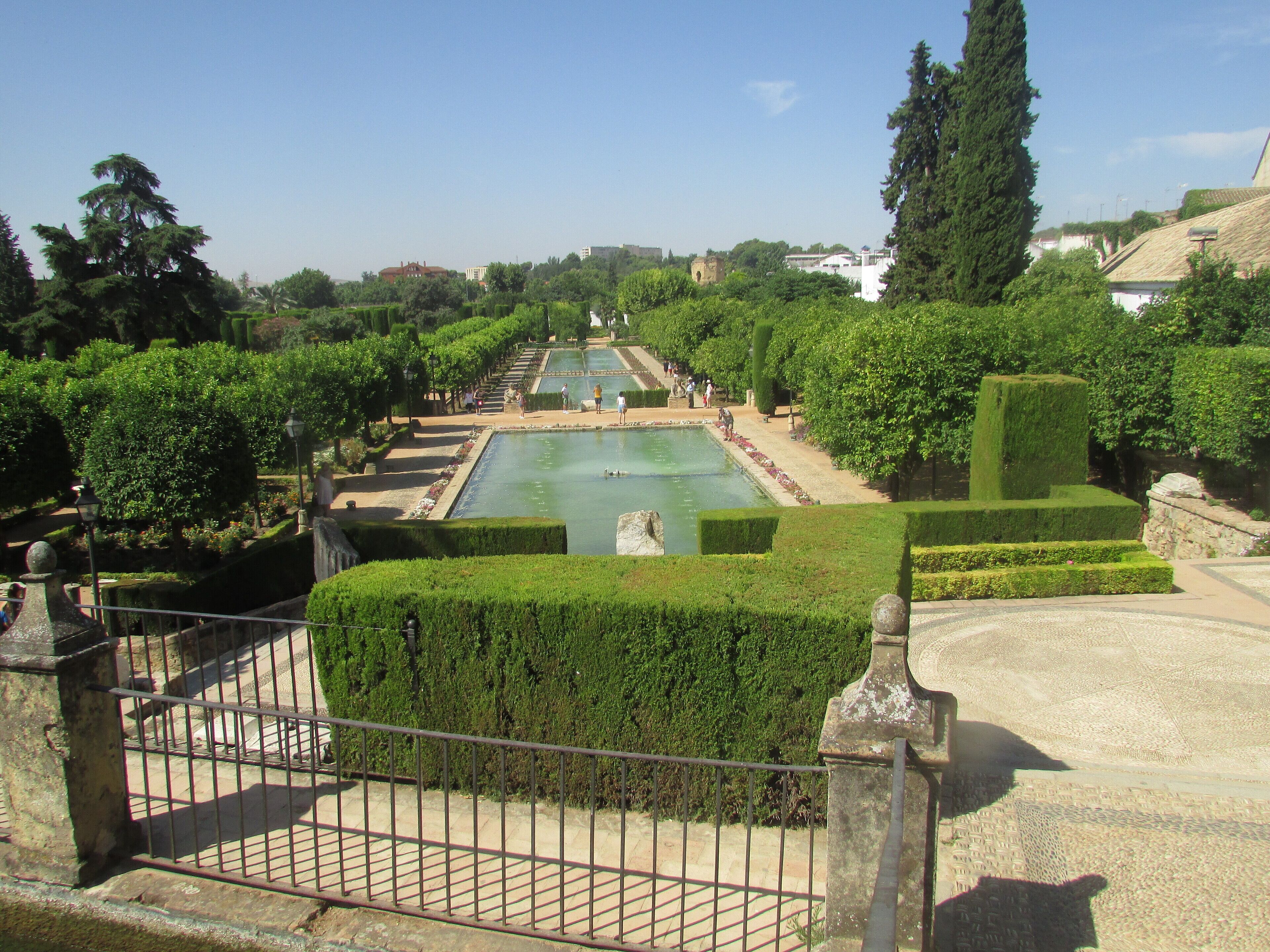 A formal garden with a central pool, neatly trimmed hedges, and a distant view of buildings.