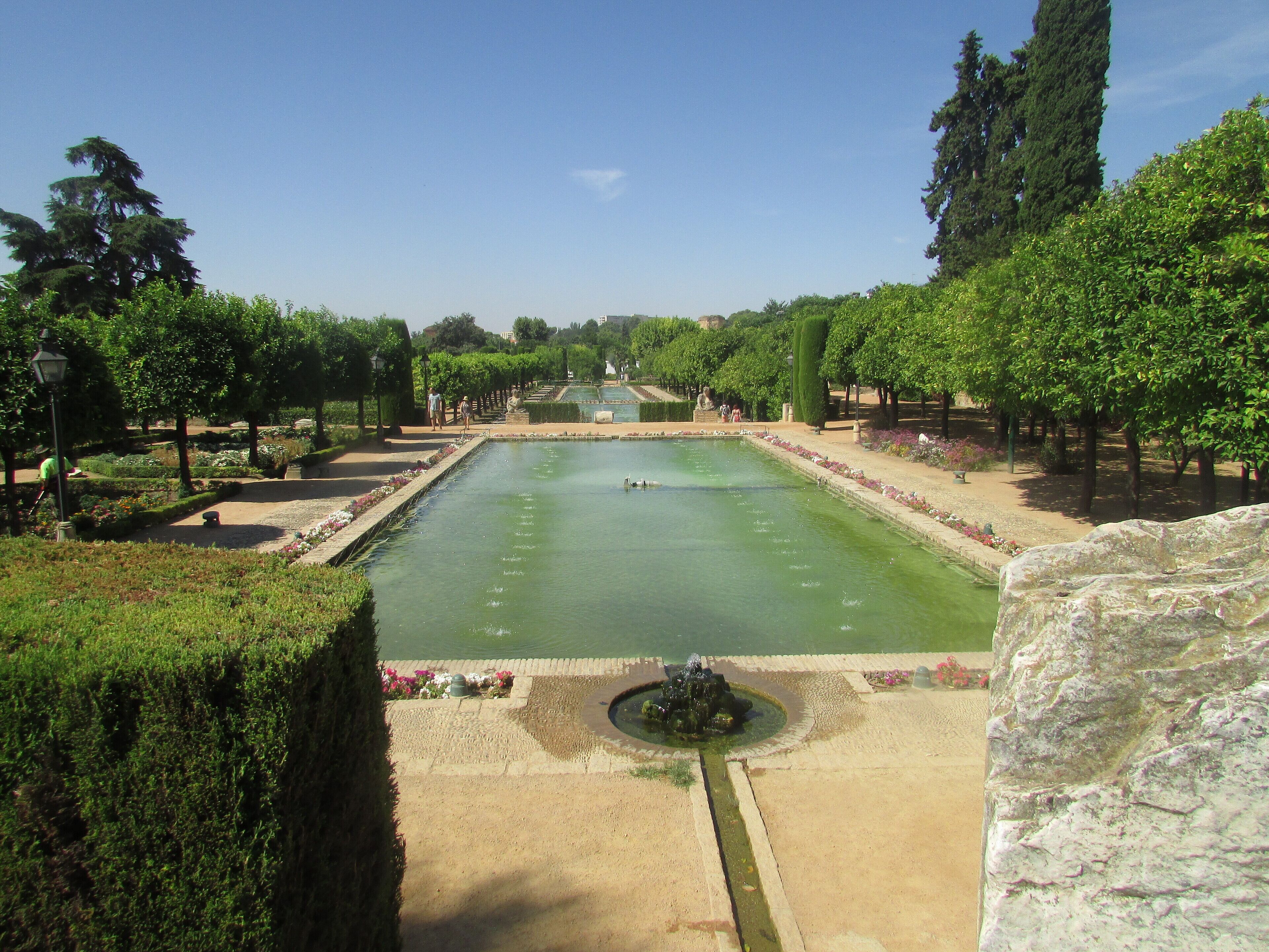 A large rectangular pool with a fountain in the center, surrounded by neatly trimmed hedges and trees.