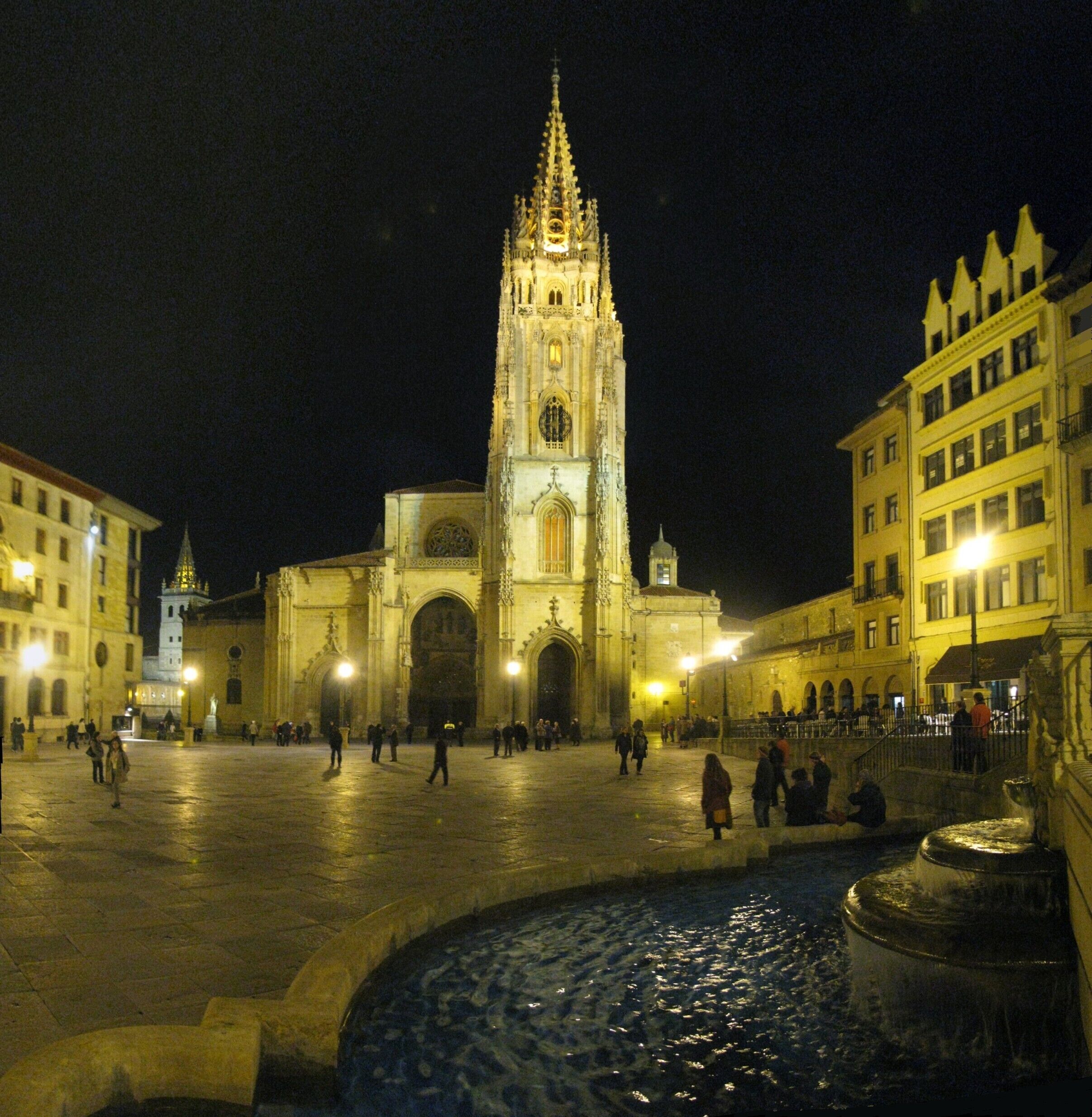 A historic cathedral with a tall spire, a fountain, and surrounding buildings at night.