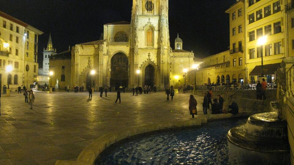A historic cathedral with a tall spire, a fountain, and surrounding buildings at night.