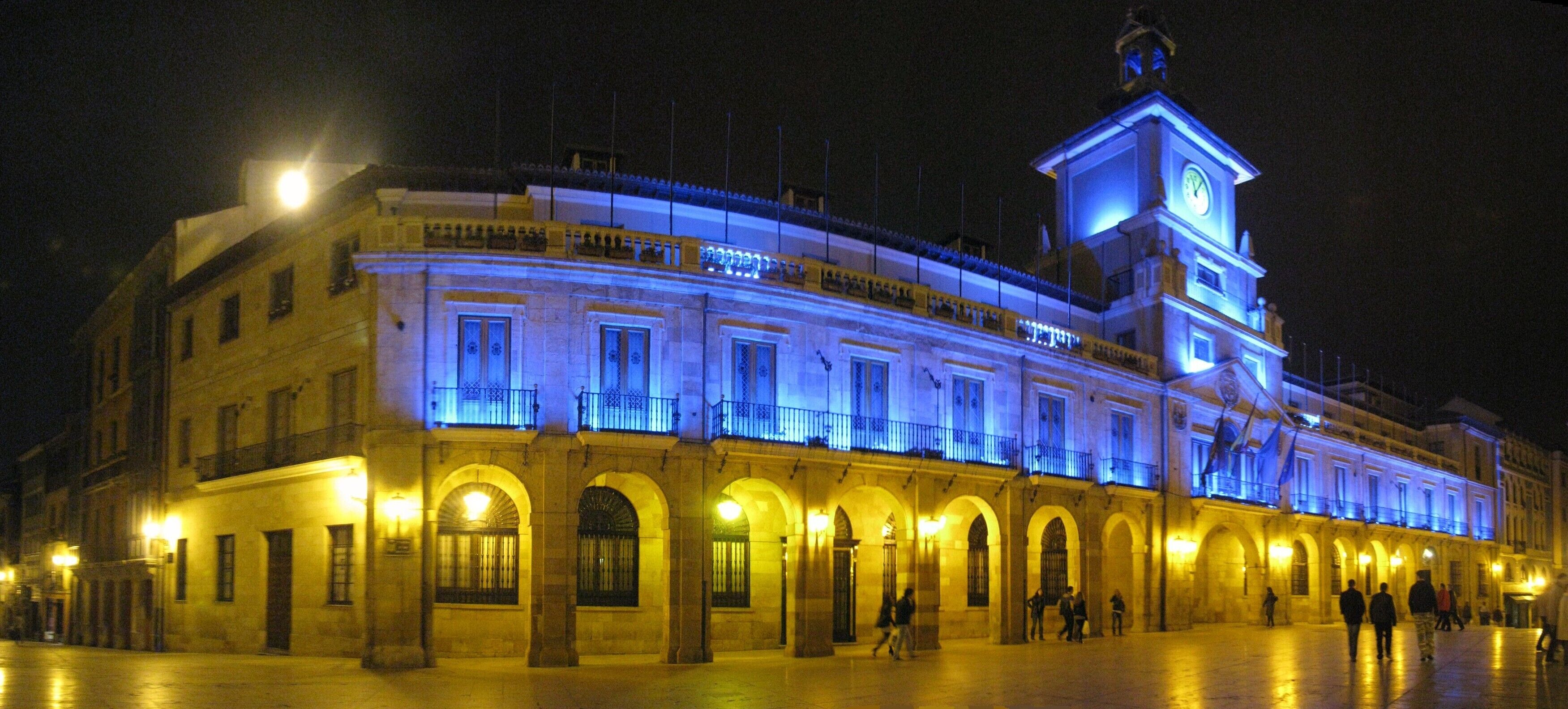 Un edificio histórico con luces azules y amarillas iluminando su fachada, arcos y una torre con reloj.