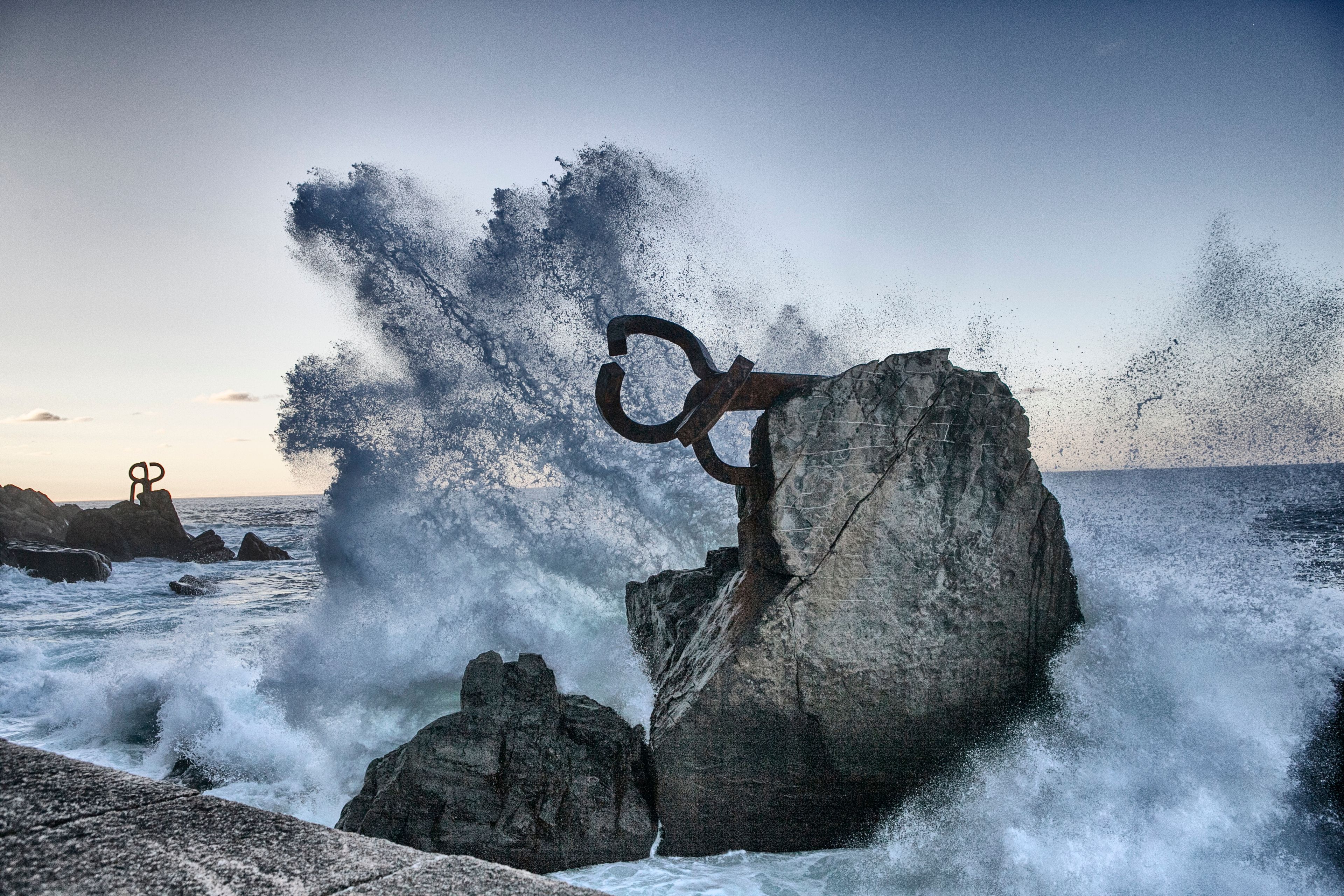 A sculpture of a figure with a circular object, set against a backdrop of crashing waves and rocky coastline.