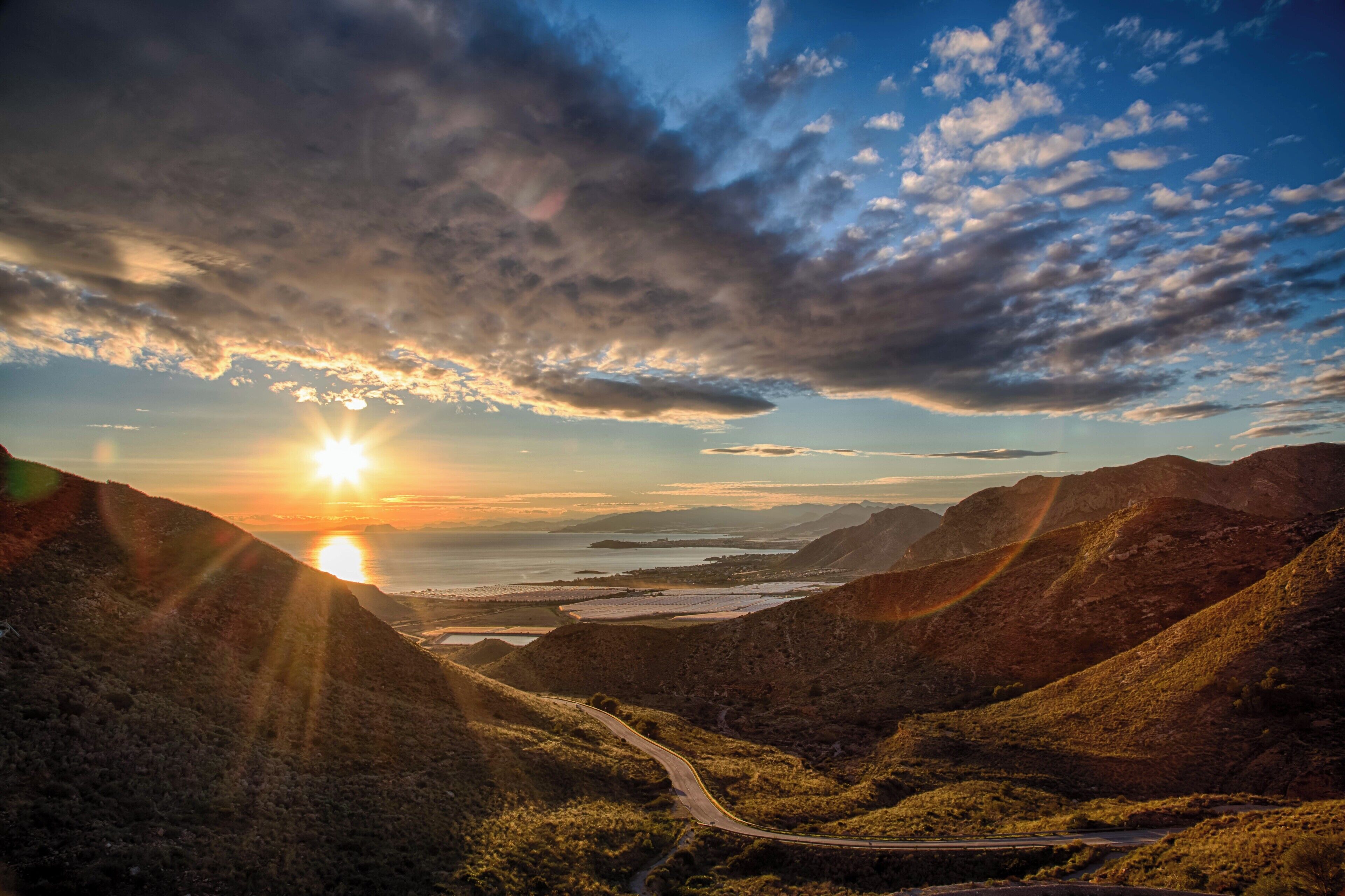 A winding road through a mountainous landscape with the sun setting behind the hills.