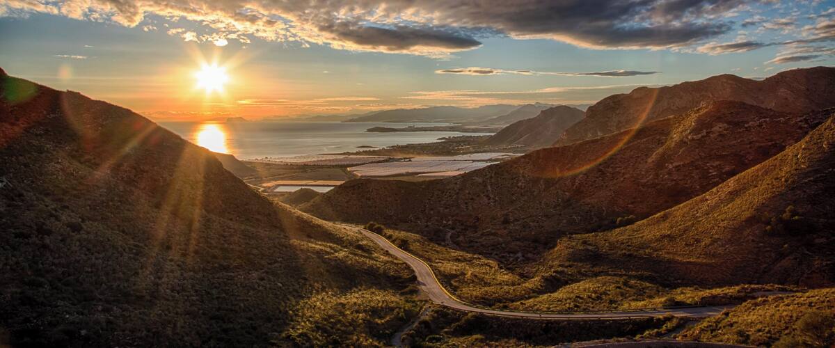 A winding road through a mountainous landscape with the sun setting behind the hills.
