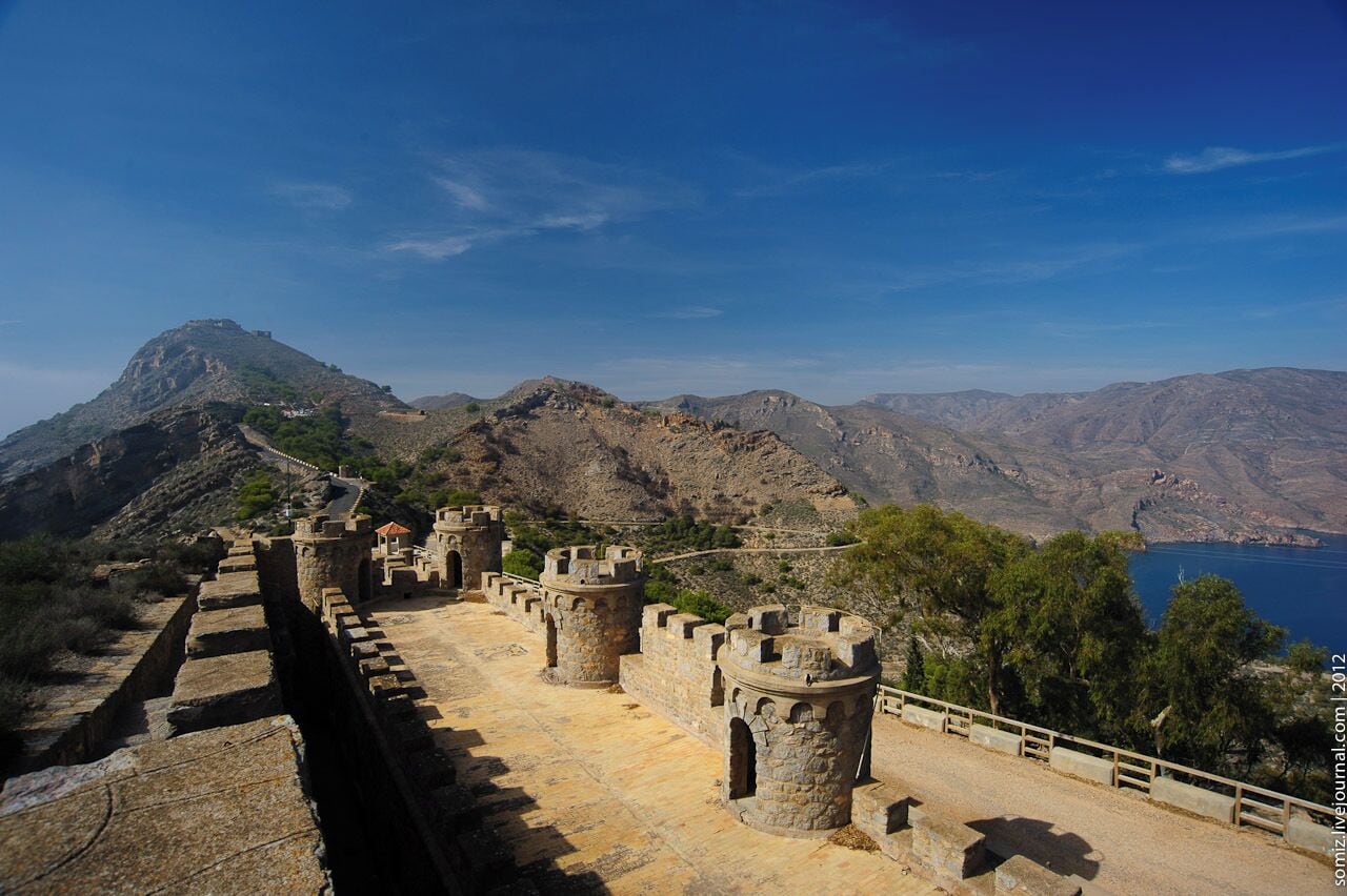 Muralha de pedra histórica com torres, com vista para uma paisagem montanhosa e um corpo de água.