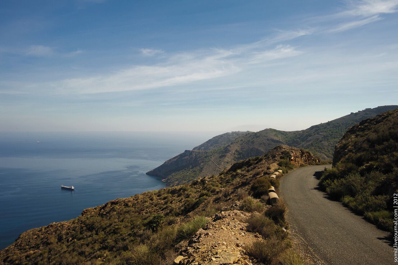 A coastal road winding through hilly terrain with a view of the sea and a distant ship.