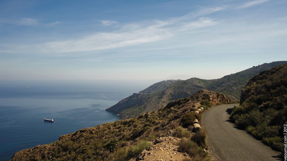 A coastal road winding through hilly terrain with a view of the sea and a distant ship.