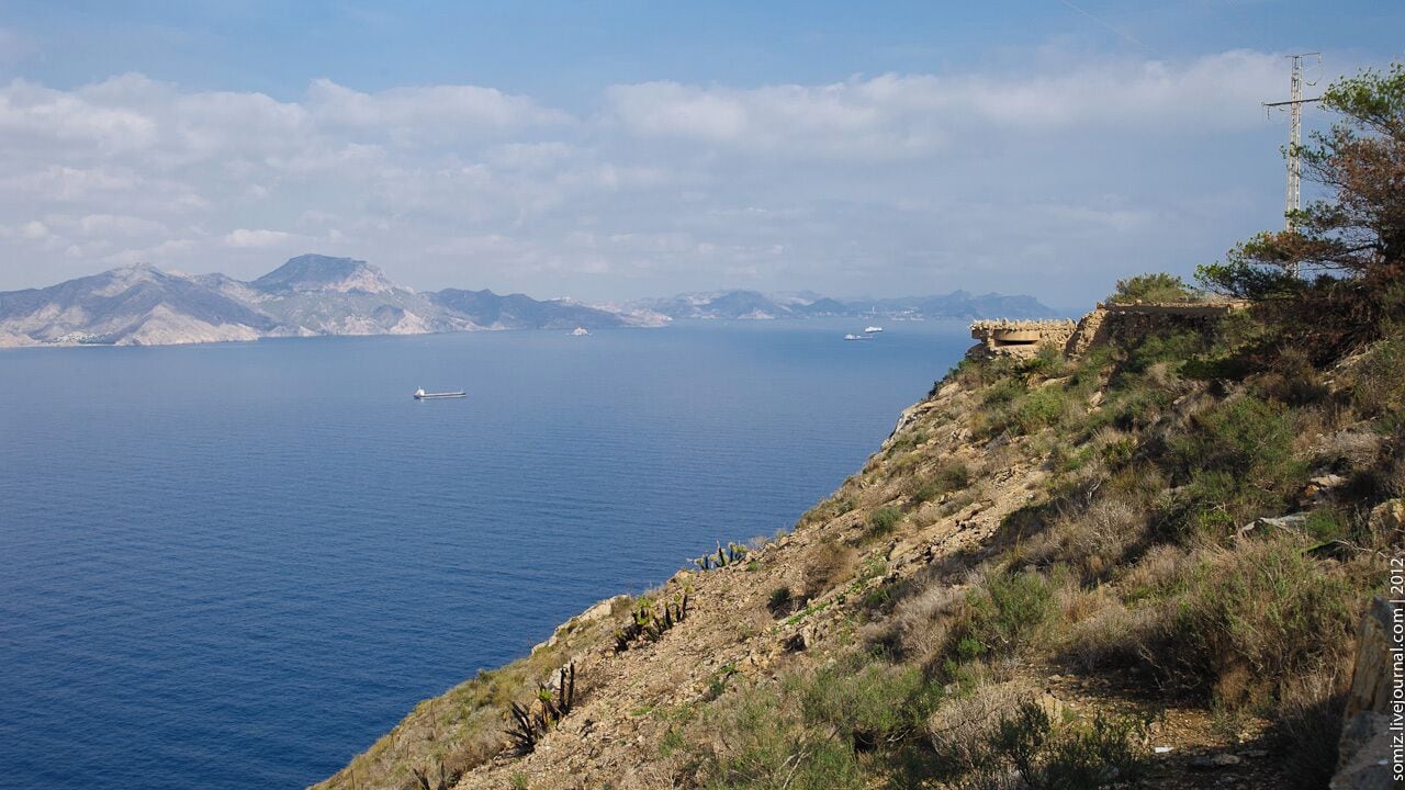 Paisagem costeira com vista nítida para o mar, um barco e montanhas ao longe.
