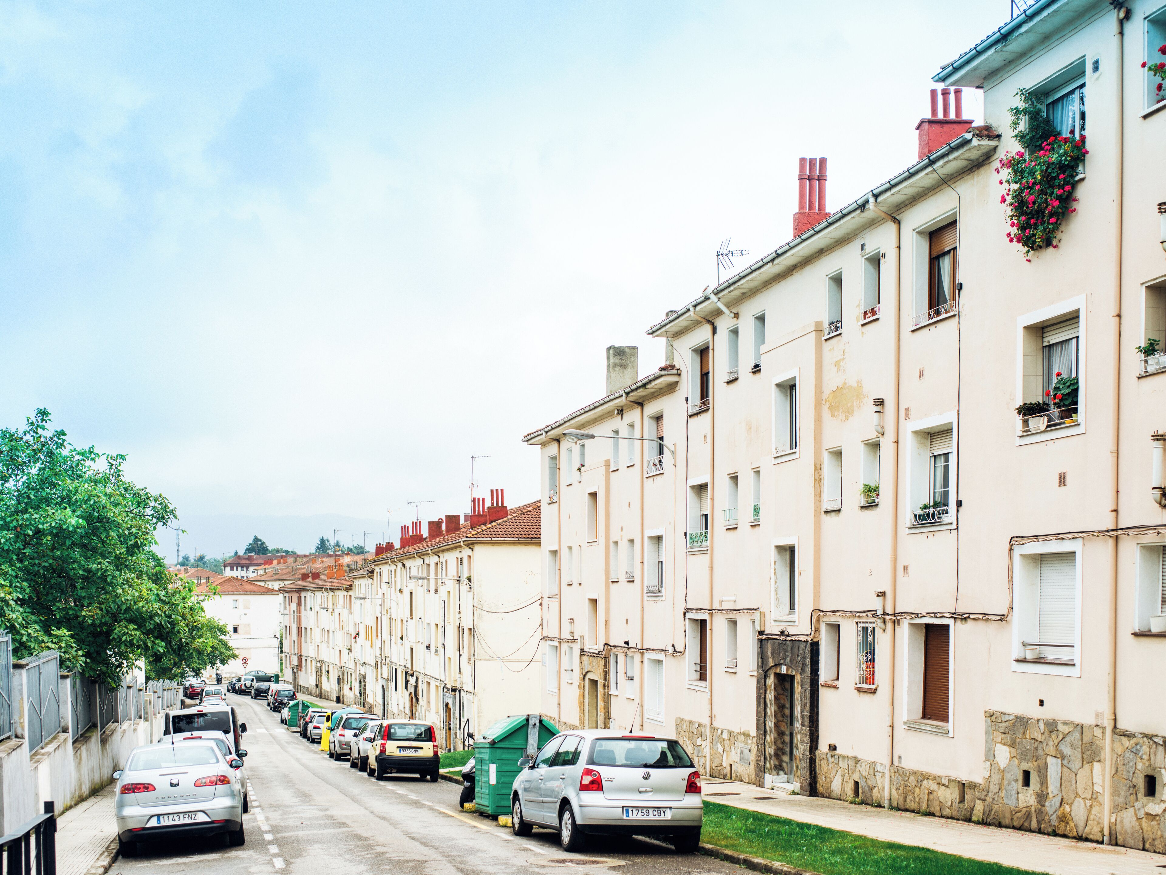 A row of parked cars along a narrow street lined with multi-story buildings, some with balconies and flower boxes.