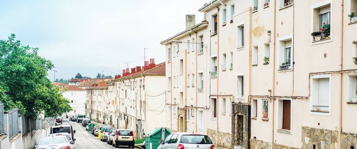 A row of parked cars along a narrow street lined with multi-story buildings, some with balconies and flower boxes.