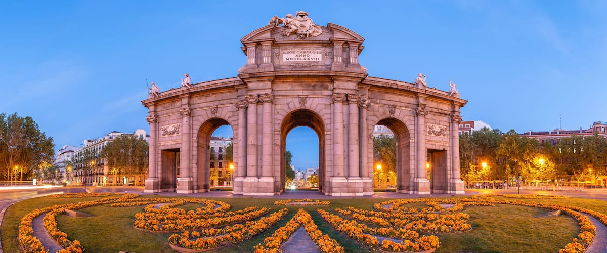 A historic archway with statues and a plaque, surrounded by a circular flower garden.