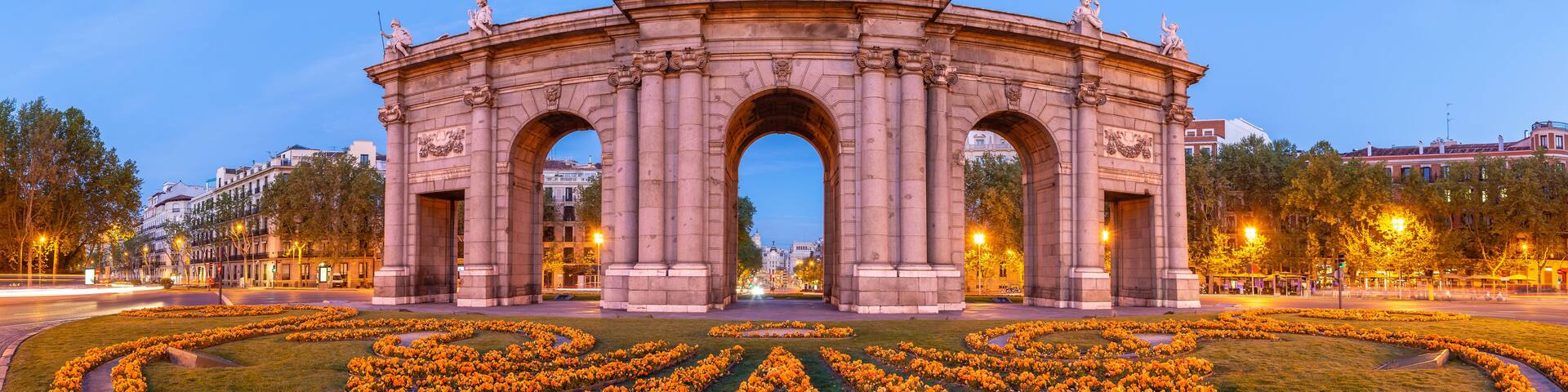 A historic archway with statues and a plaque, surrounded by a circular flower garden.
